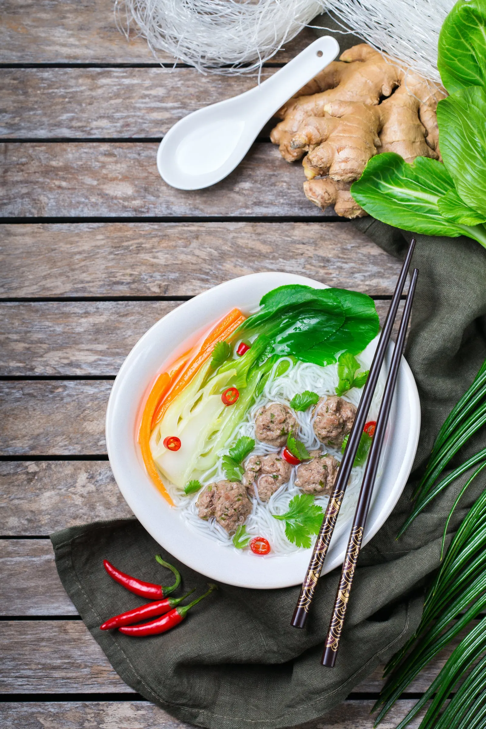 A bowl of rice vermicelli with pork meatballs, pak choi and coriander on a wooden table, with ginger and red chillies alongside