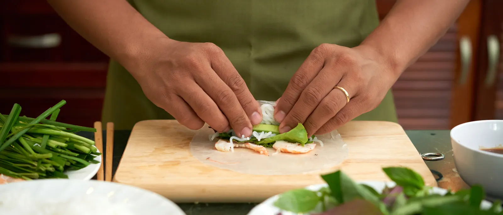 Hands rolling a fresh summer roll at a prep board, with herbs, rice noodles and dipping sauce set out around it